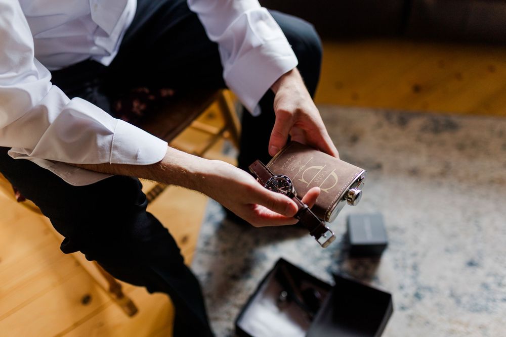 Man in white shirt holding an engraved brown leather flask while seated, with black gift boxes on the floor.