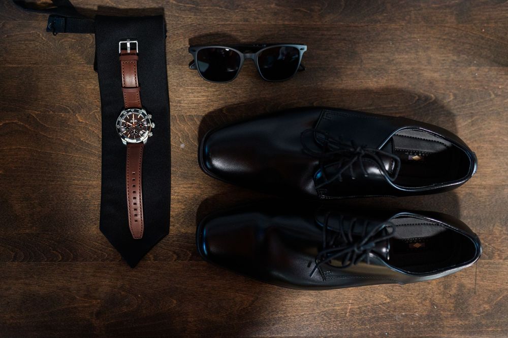Black dress shoes, tie, watch, and sunglasses arranged on a wooden surface for a formal mens accessory flat lay.