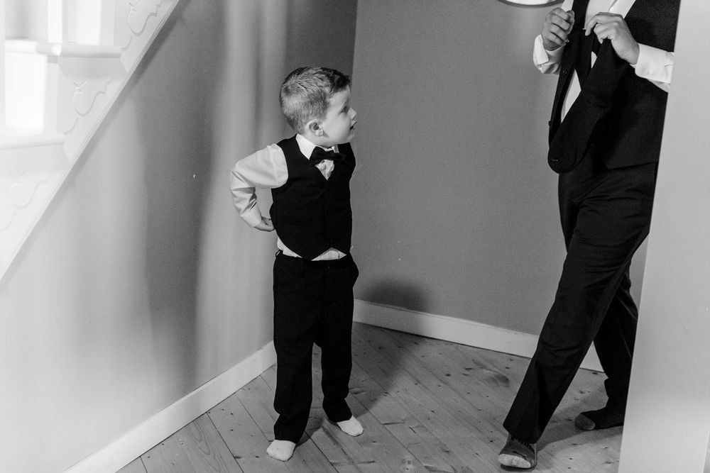 Black and white photo of a young ring bearer in a vest and bow tie standing beside a man in a tuxedo.