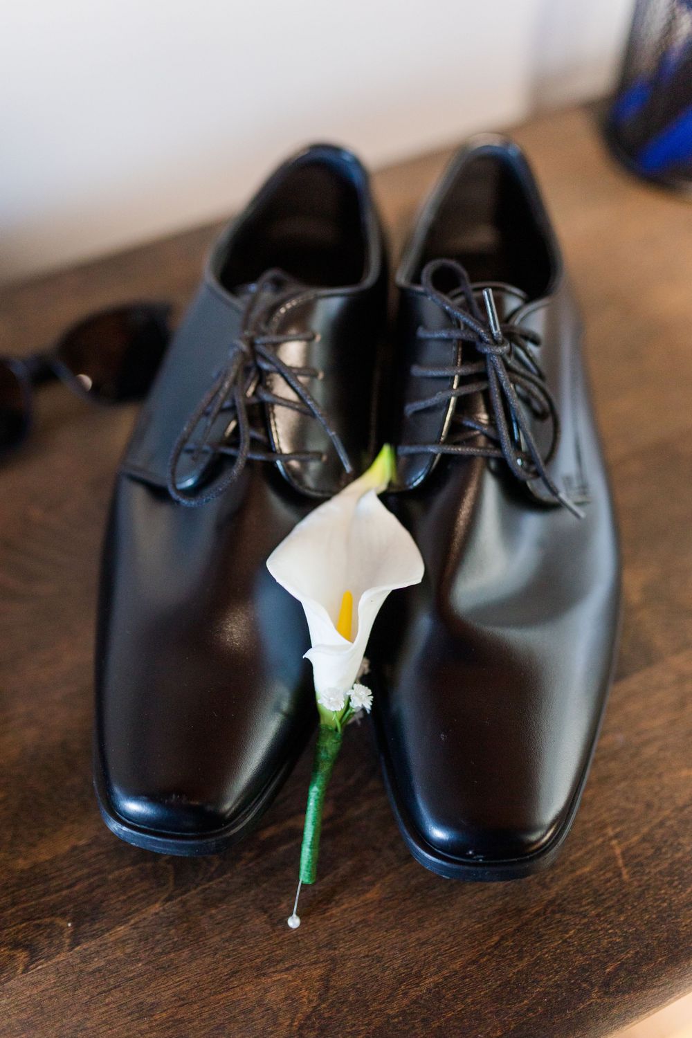 Black leather dress shoes with laces and a white calla lily boutonniere on a wooden surface.