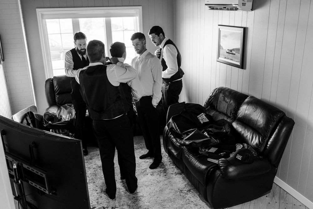 Groomsmen helping each other prepare in a room before a wedding ceremony, black and white photo.