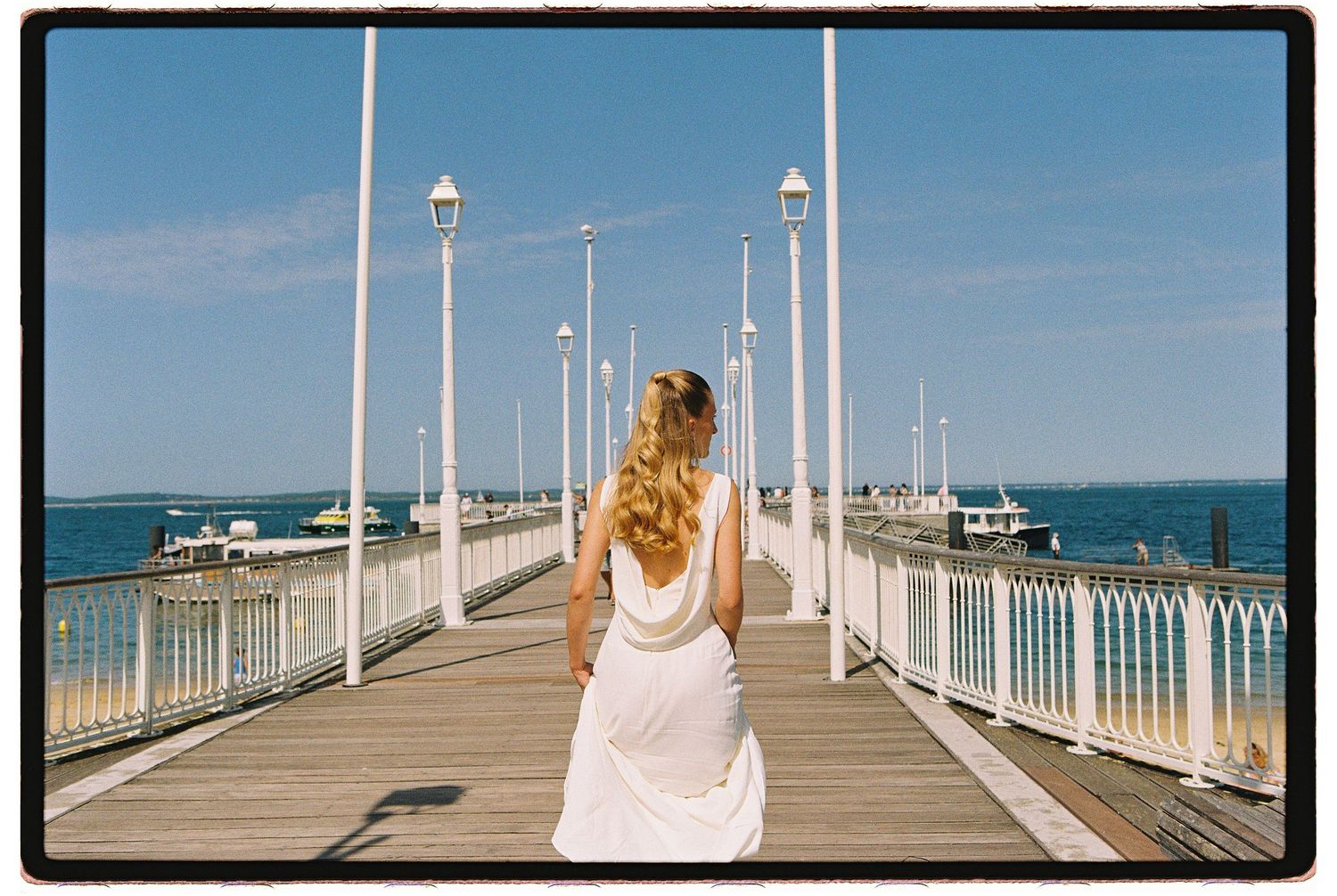 A woman in a white dress walks along a sunlit pier with white lamp posts and boats in the background.