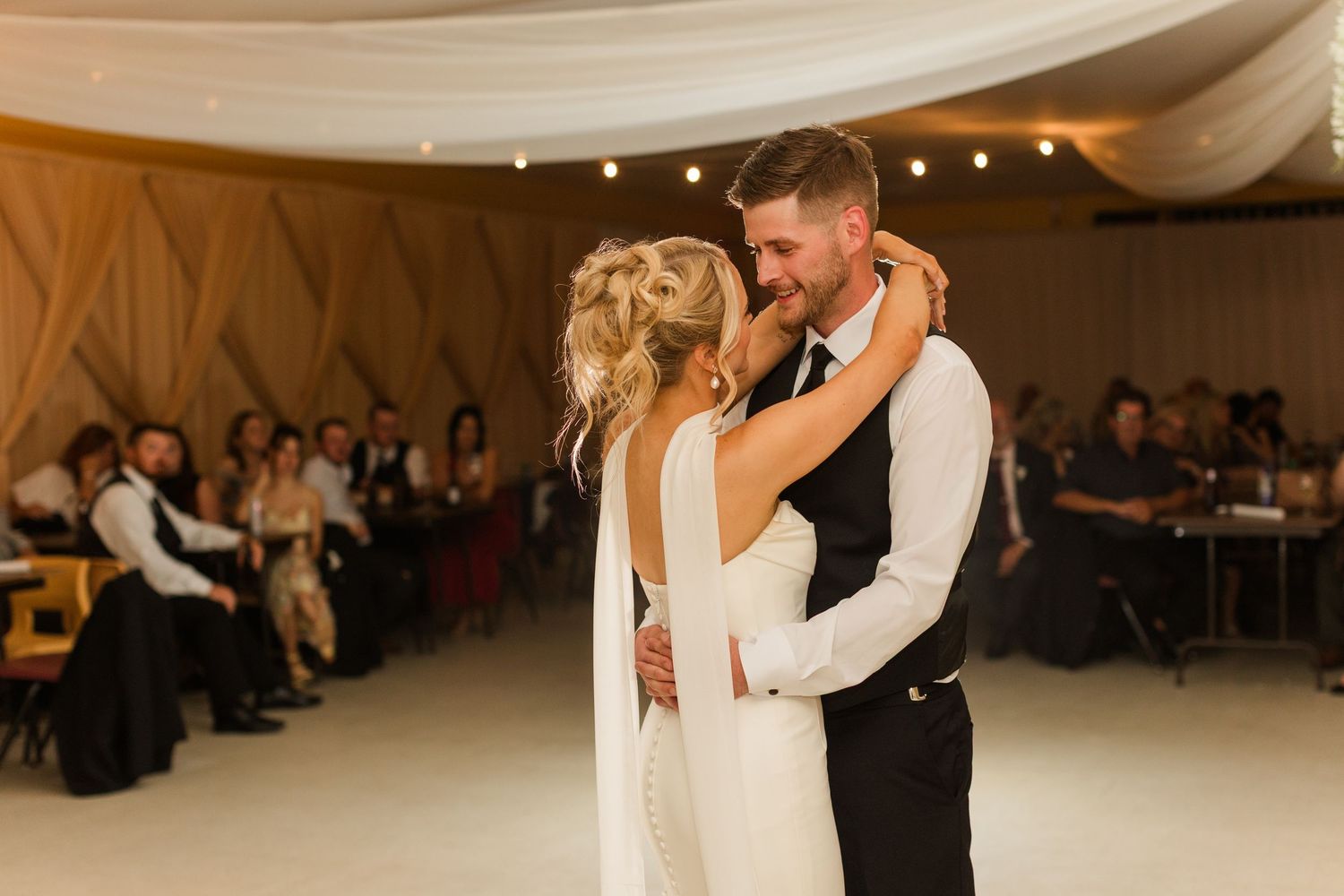 Bride and groom share their first dance at a wedding reception, surrounded by seated guests in a warmly lit venue.
