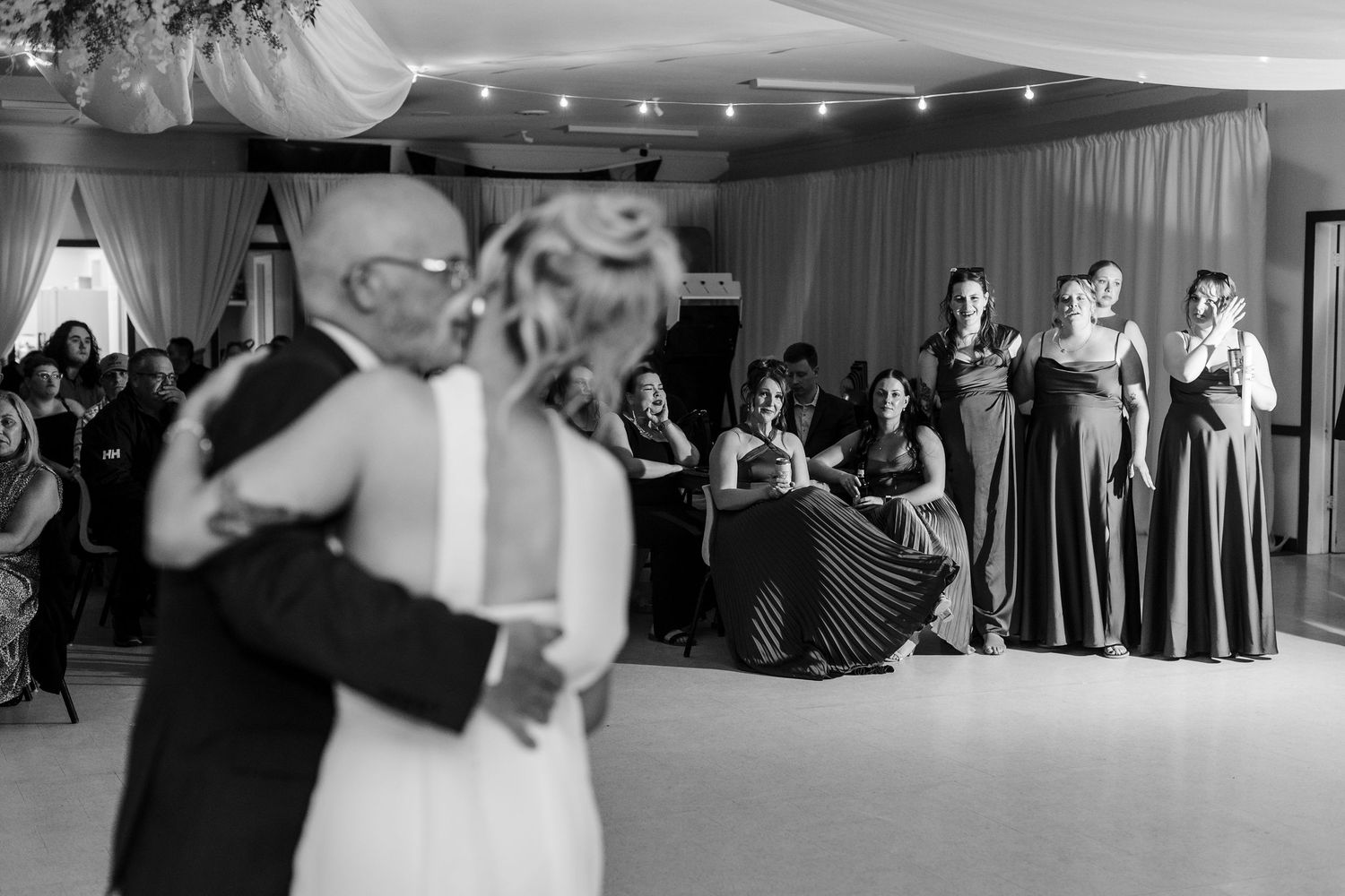 Black and white wedding photo of couple sharing first dance while bridesmaids watch at elegant reception venue.