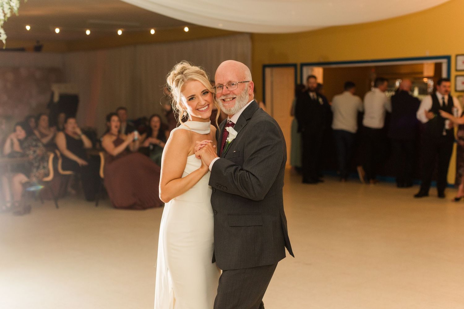 Bride and father share a tender first dance at wedding reception, guests watching in background.