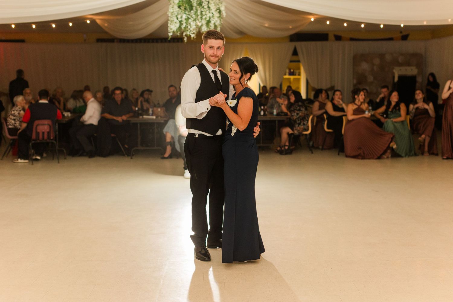 Couple sharing a first dance on white dance floor at elegant wedding reception with guests seated in background.