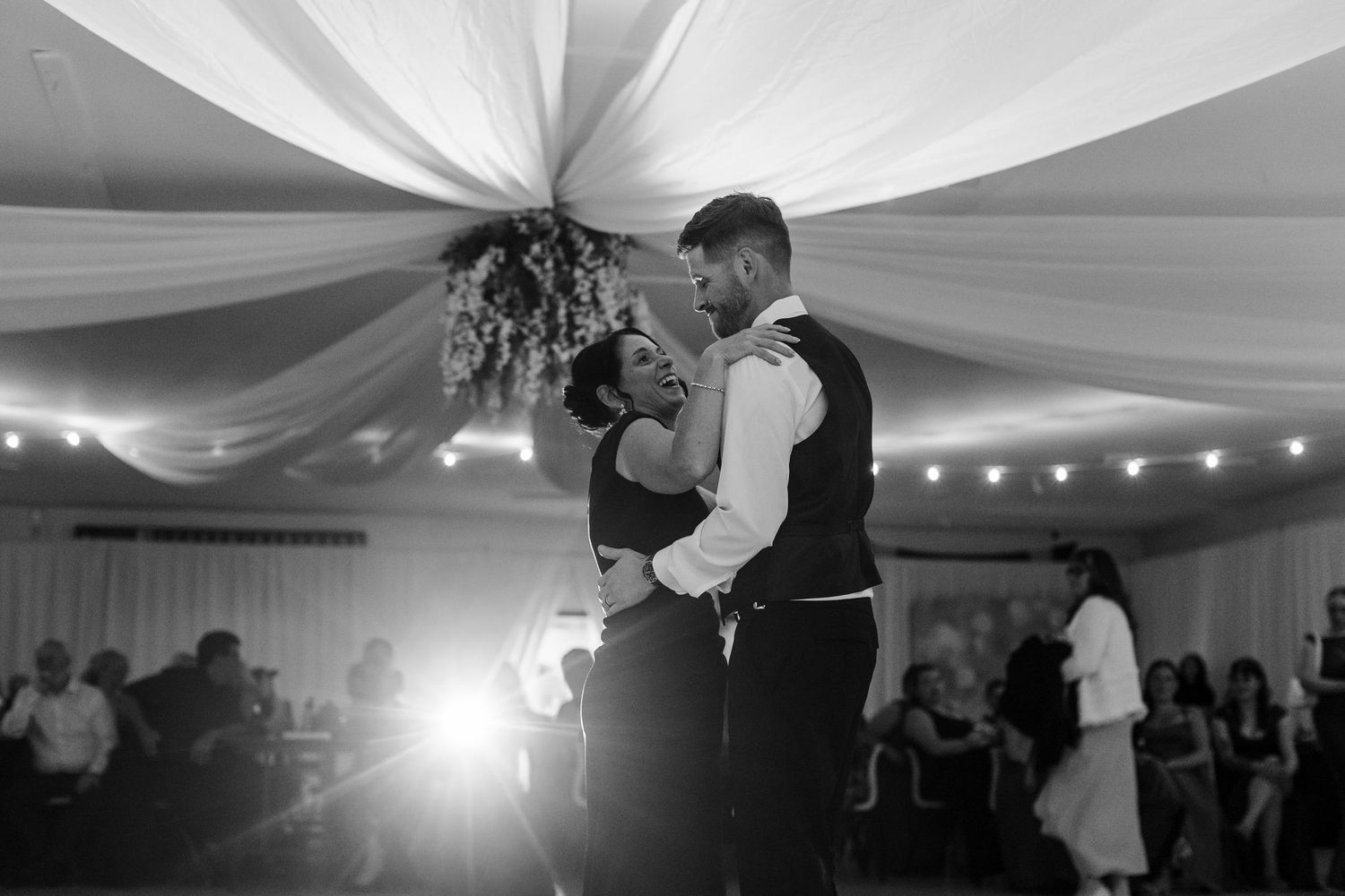 Black and white photo of a couple sharing their first dance at a wedding reception under draped fabric ceiling.