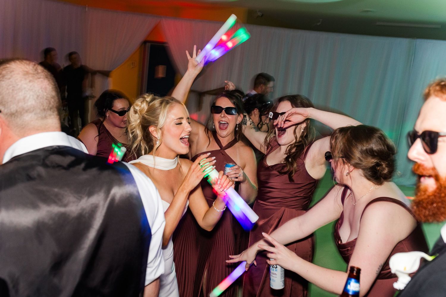 Bride and bridesmaids in burgundy dresses dancing with LED foam sticks at a lively wedding reception party.