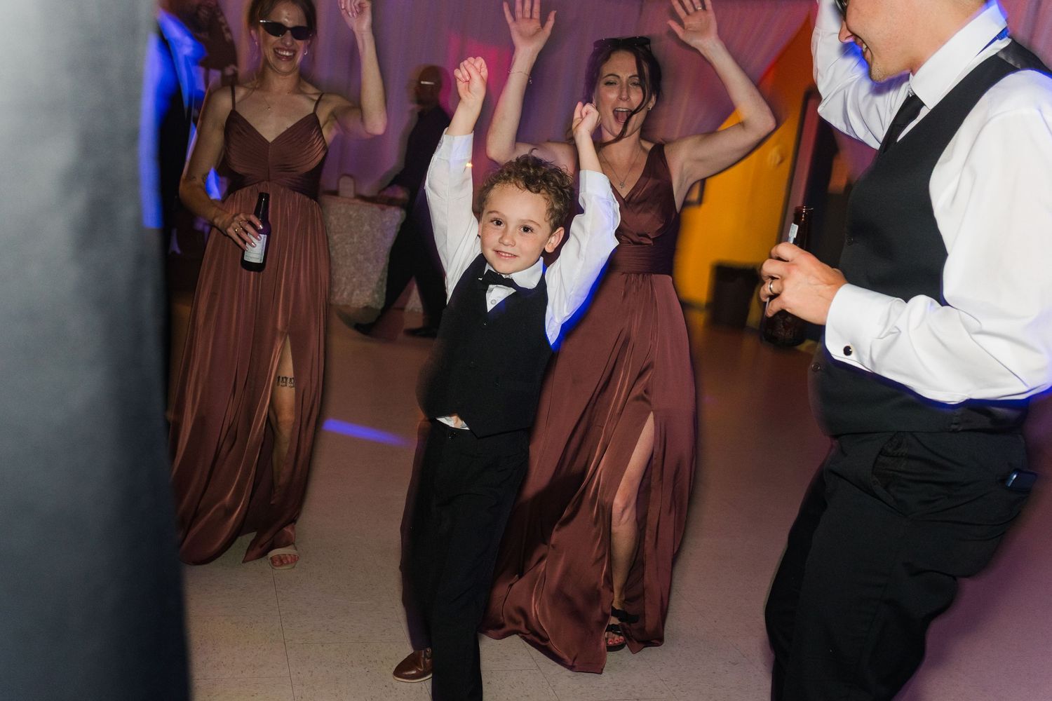 Young boy in black suit dancing joyfully at wedding reception with bridesmaids in brown gowns.
