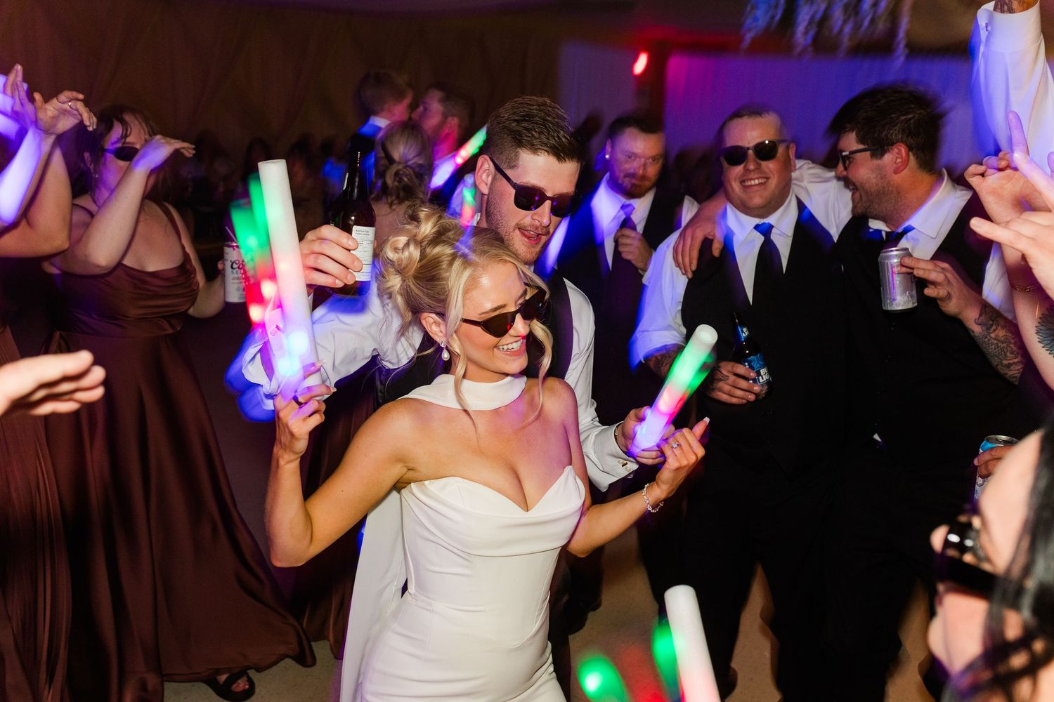 Bride and groom dancing with guests holding colorful glow sticks at a lively wedding reception party.