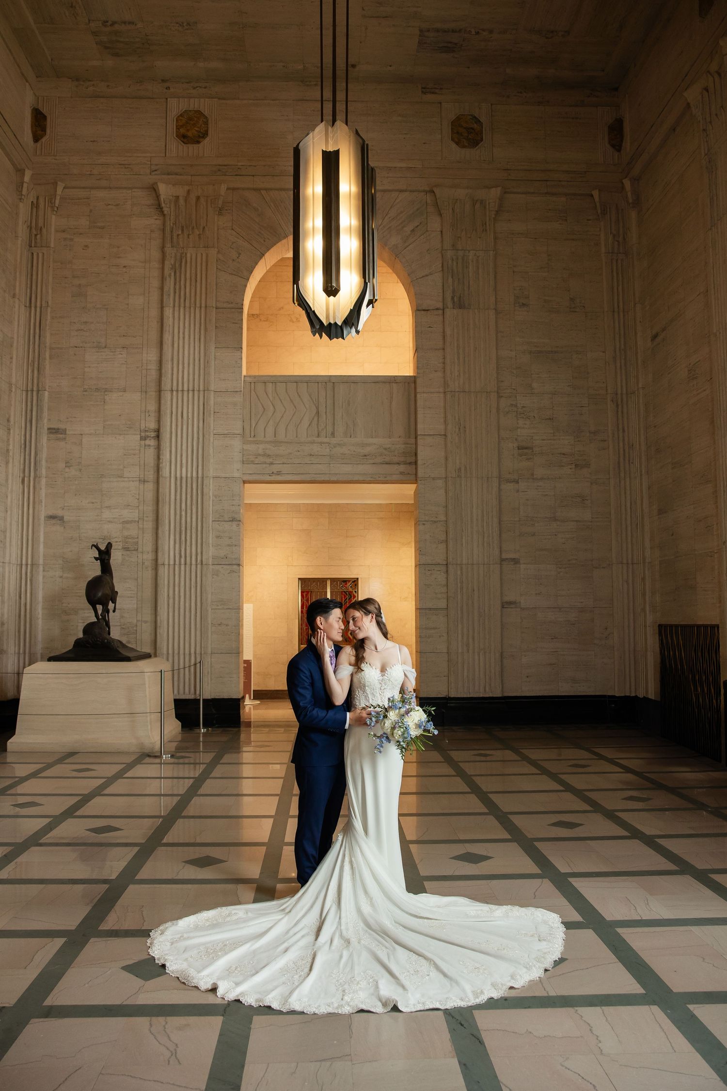 Bride and groom embrace in elegant marble hall with Art Deco chandelier, bride's long train spread across checkered floor.