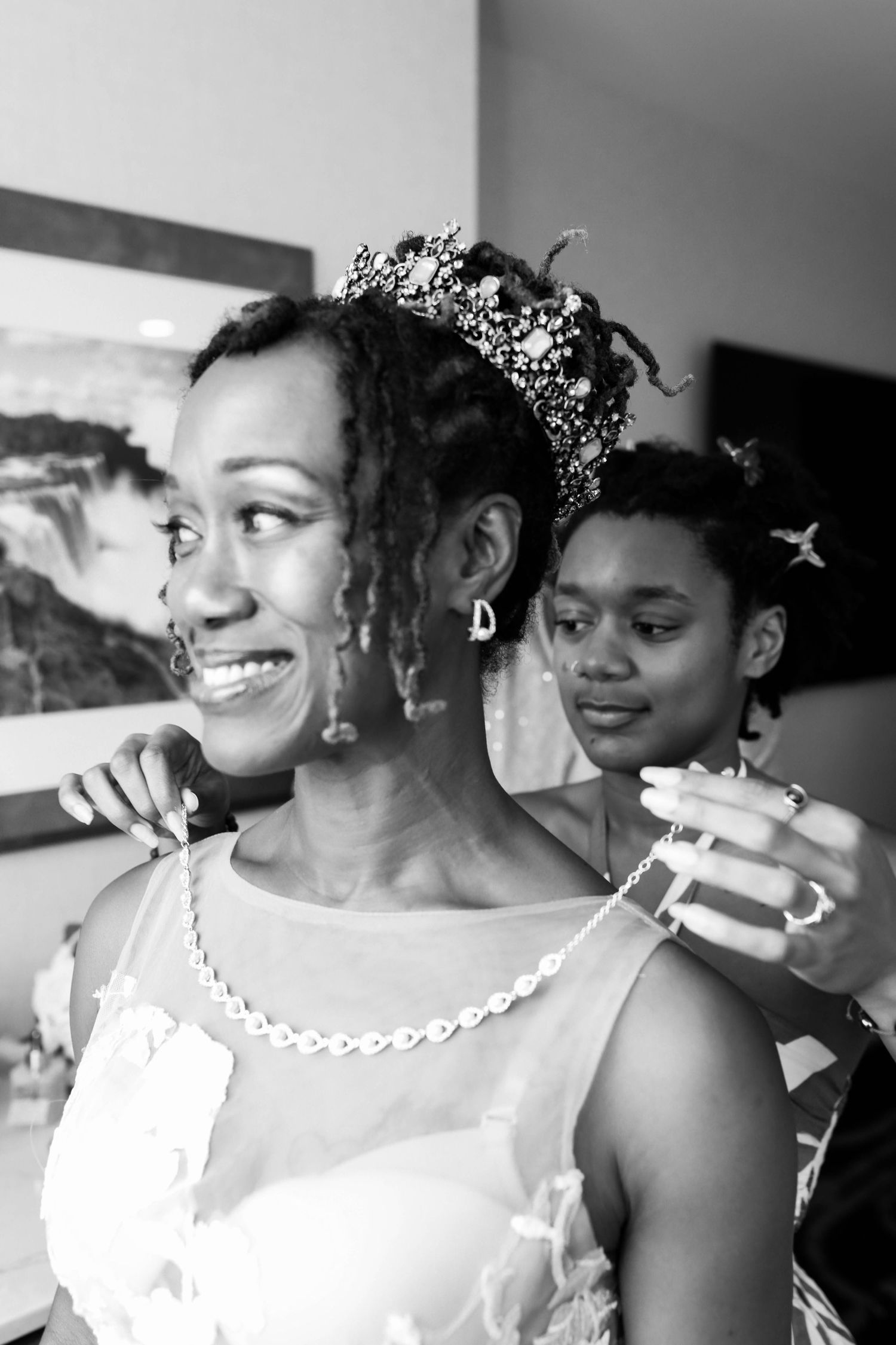 Smiling bride in a lace gown and jeweled tiara has a pearl necklace fastened by a bridesmaid in this black and white getting-ready photo.