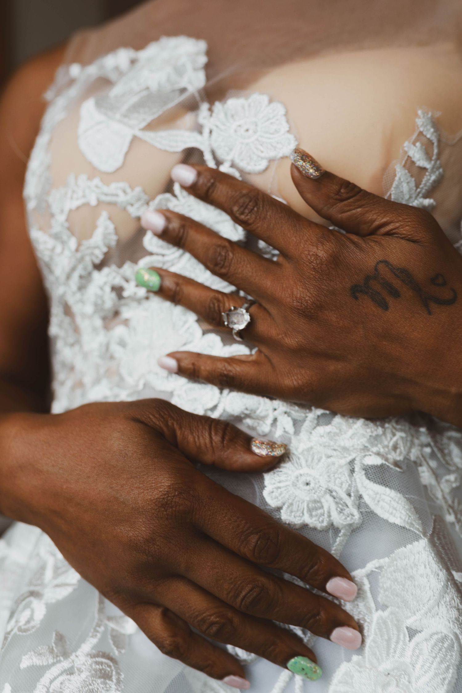 Bride's hands with pastel manicure, diamond ring and a small tattoo rest on a white floral lace wedding dress.
