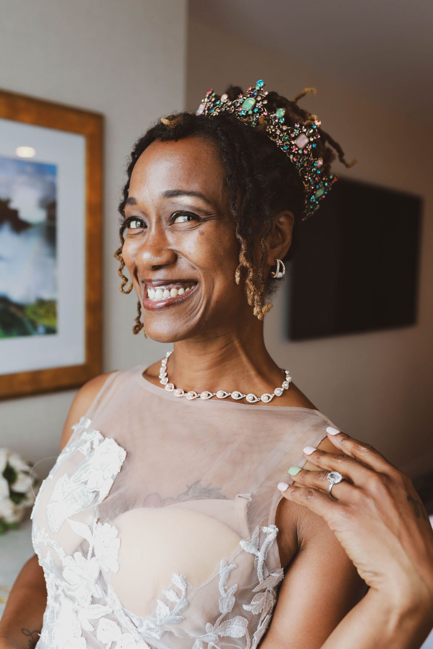 Smiling bride in a lace wedding gown wears a jeweled crown and pearl necklace while her hand rests on her shoulder.