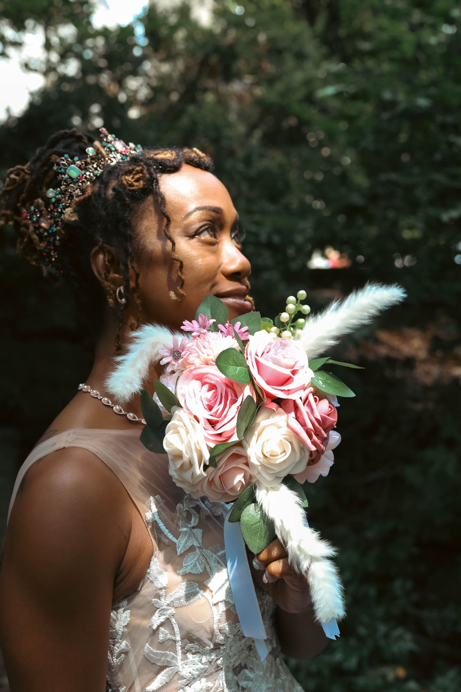 Bride in a lace gown and jeweled headpiece gazes upward while holding a pink and white rose bouquet with feather accents outdoors.