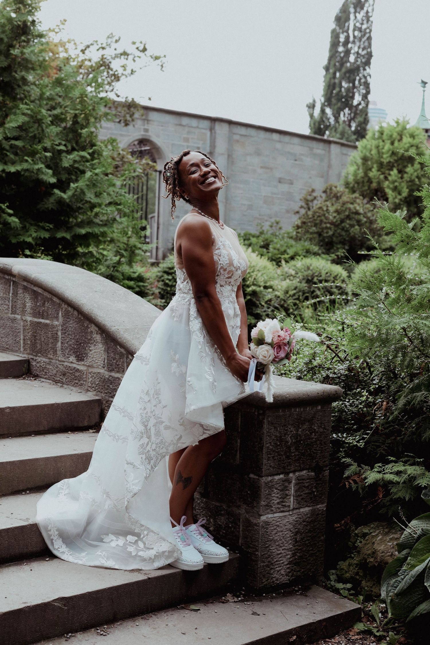 A smiling bride in a lace wedding dress and white sneakers poses on stone garden steps, holding a pink and white bouquet.
