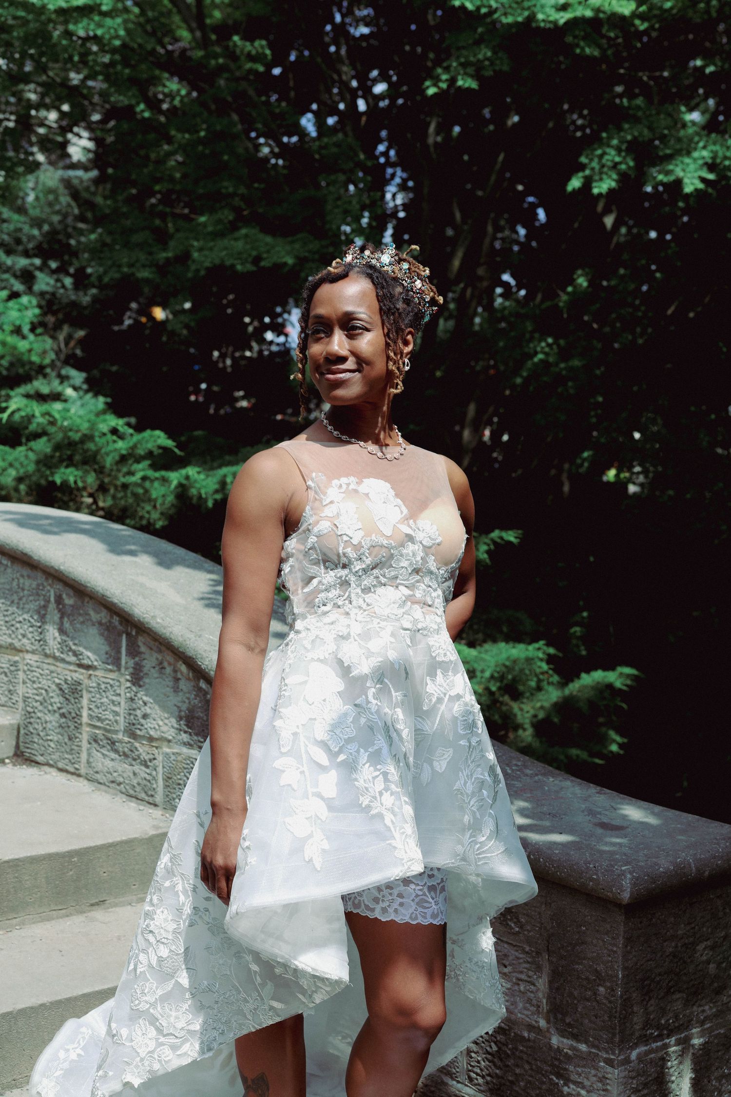 Smiling bride in a white floral lace high-low wedding dress and floral hair crown stands beside a stone staircase surrounded by green trees.