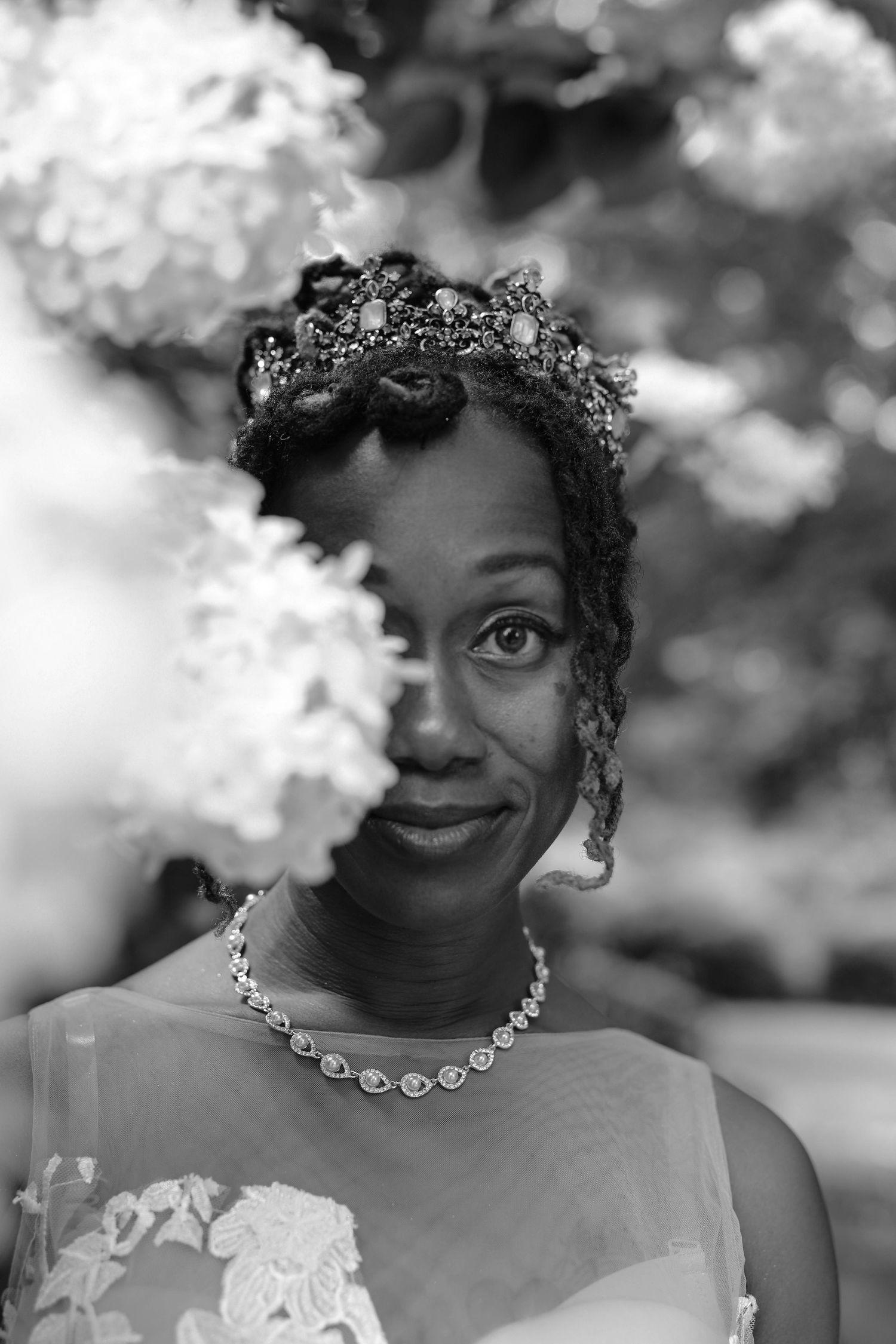 Black and white portrait of a smiling bride in a jeweled tiara and crystal necklace peeking from behind white hydrangea blooms.