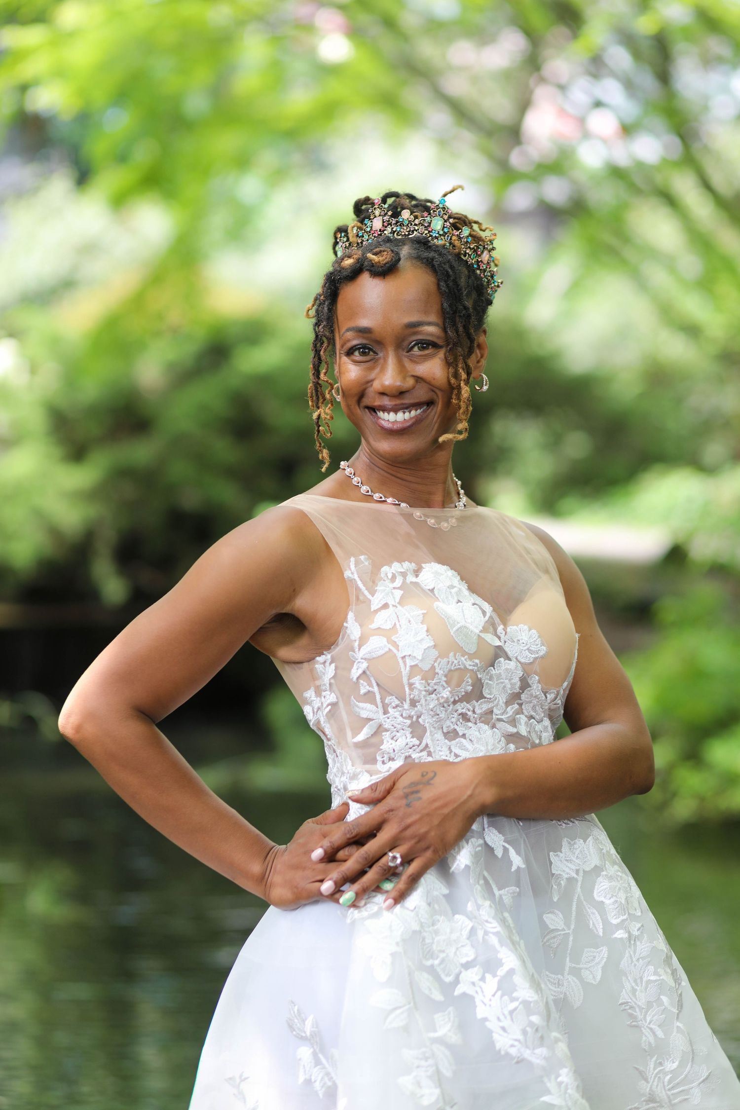 Smiling bride in a white floral lace gown and beaded tiara poses with hands on hips beside a tranquil pond in a lush green garden.