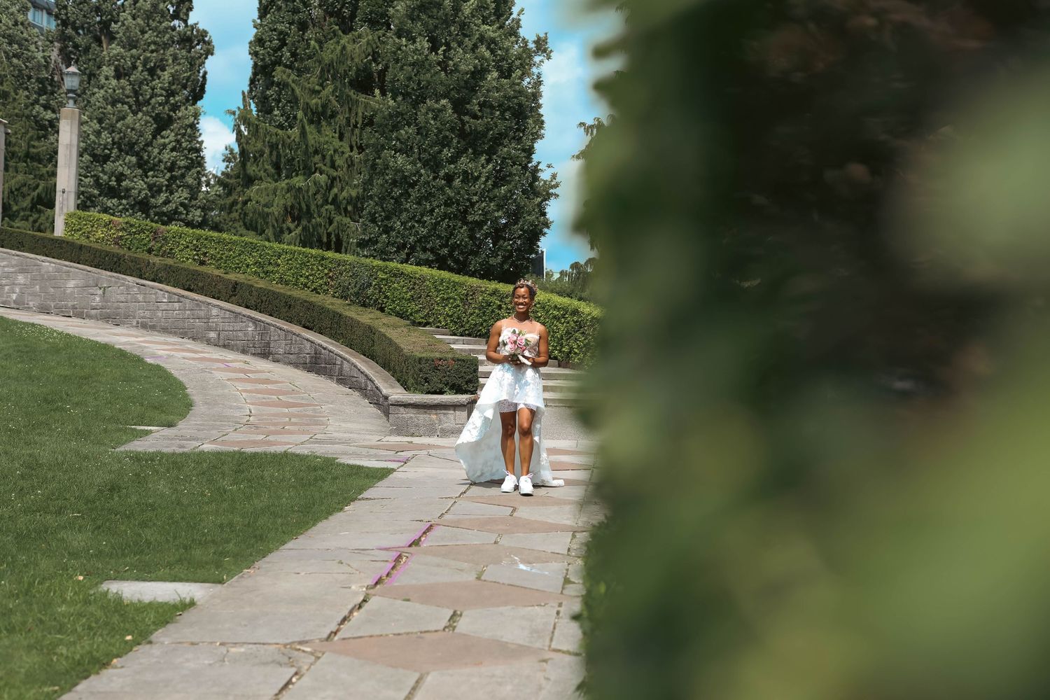 A bride in a white wedding dress walks along a stone garden path lined with manicured hedges and tall evergreen trees on a sunny day.