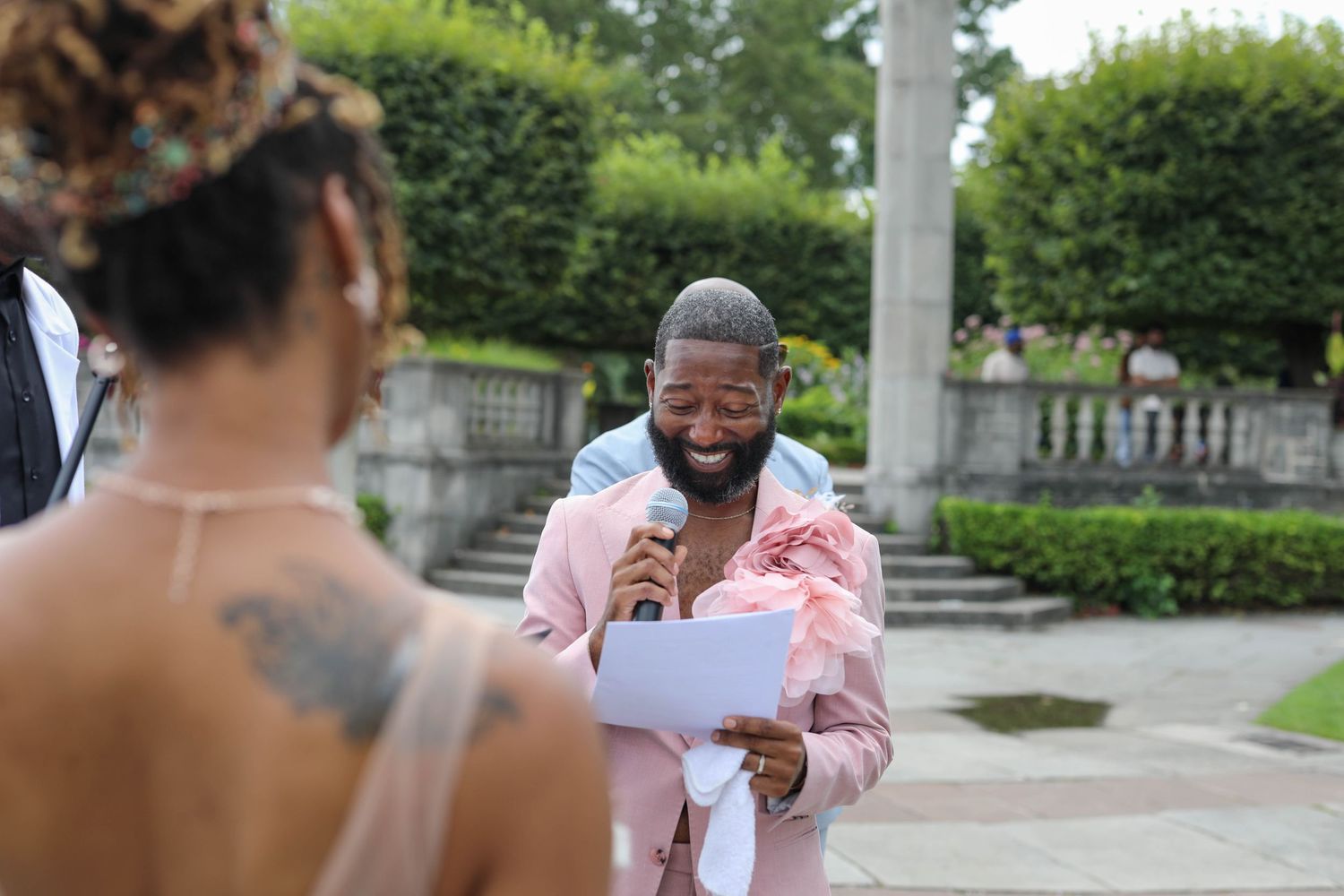 Smiling man in a pink suit reads from paper into a microphone during an outdoor garden ceremony while a woman with a back tattoo watches.