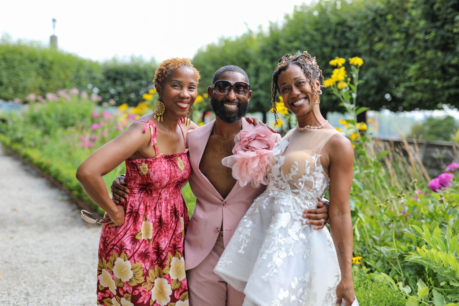 Three smiling people embrace in a sunlit garden, a woman in a floral dress, a man in a pink suit, and a woman in a white lace gown.
