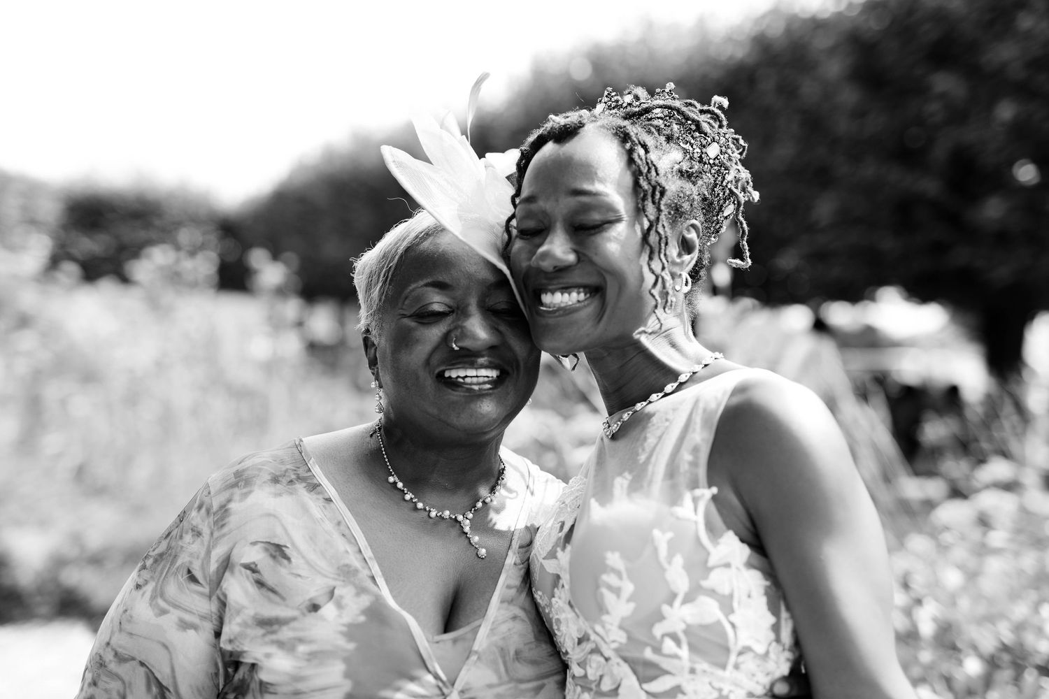 Two smiling women in floral dresses embrace cheek to cheek in a joyful black and white portrait, one wearing a fascinator and pearls.