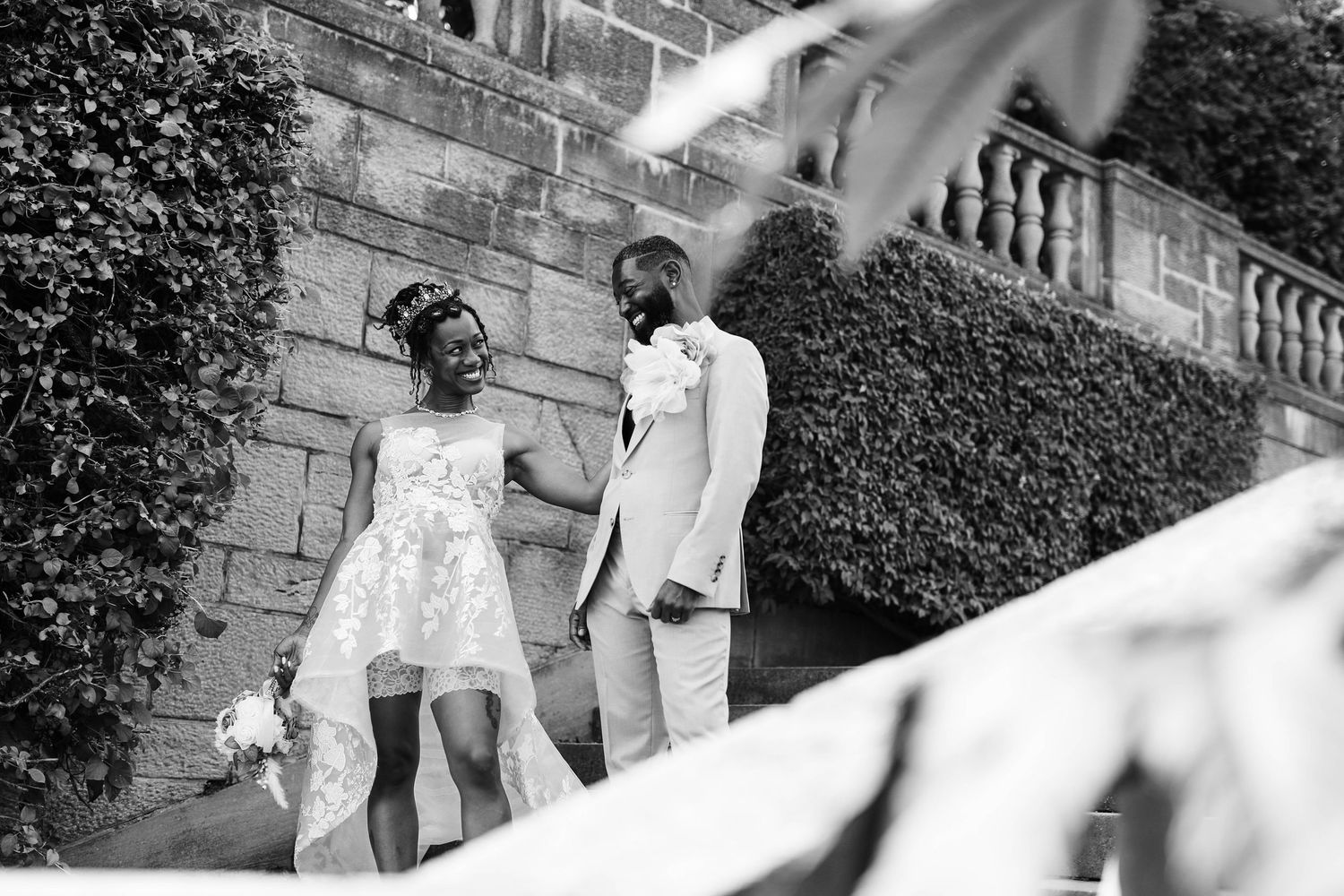 Black and white photo of a bride in a lace wedding dress and a groom in a light suit sharing a tender moment by a stone wall.