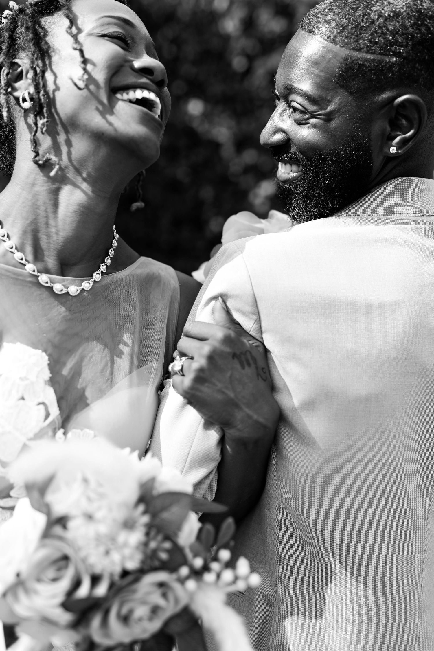 Black and white photo of a joyful bride and groom laughing together, the bride in a pearl necklace holding a bouquet of roses.