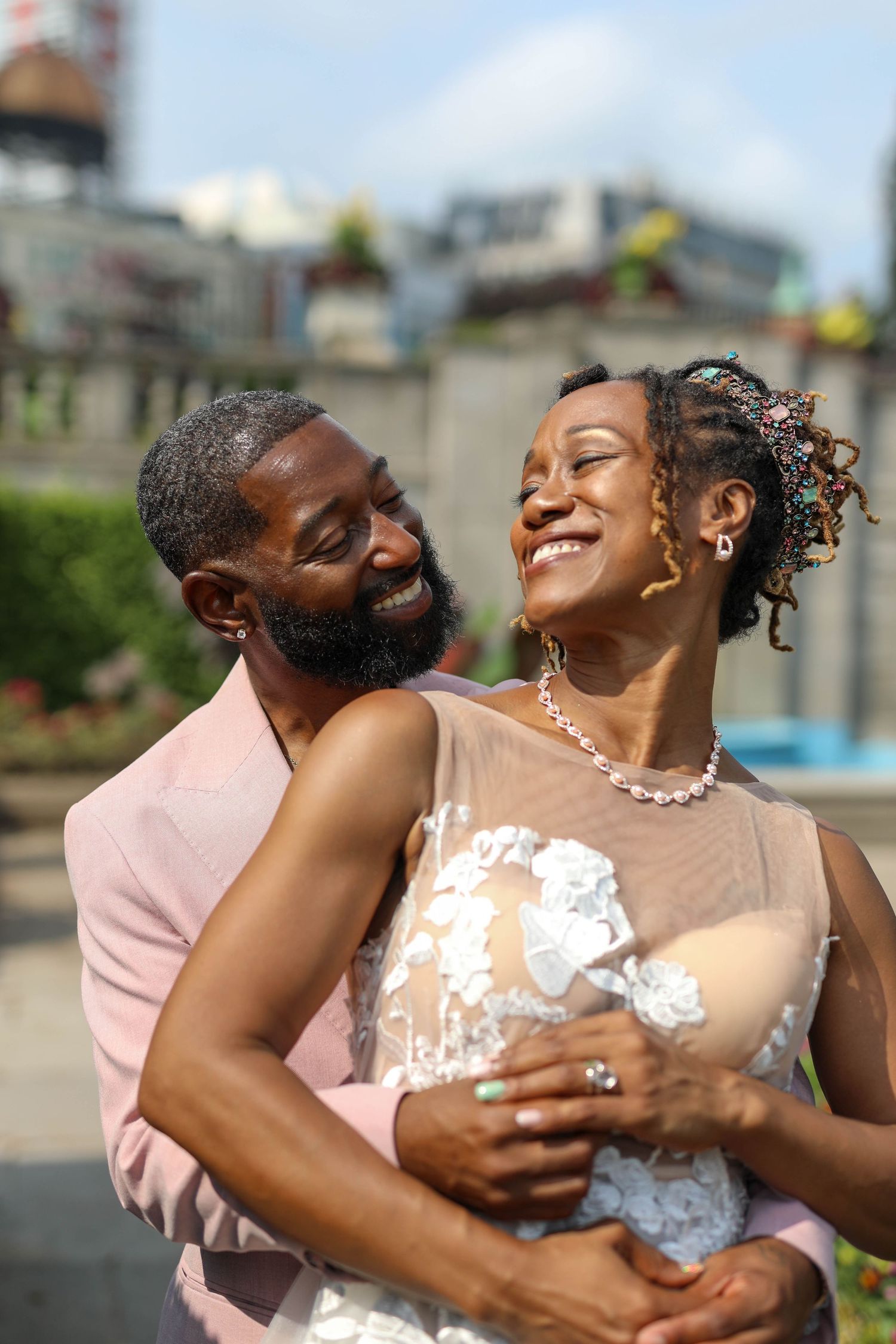 Smiling bride in a lace wedding dress and floral hair accessory embraces her groom in a pink suit during an outdoor city wedding portrait.