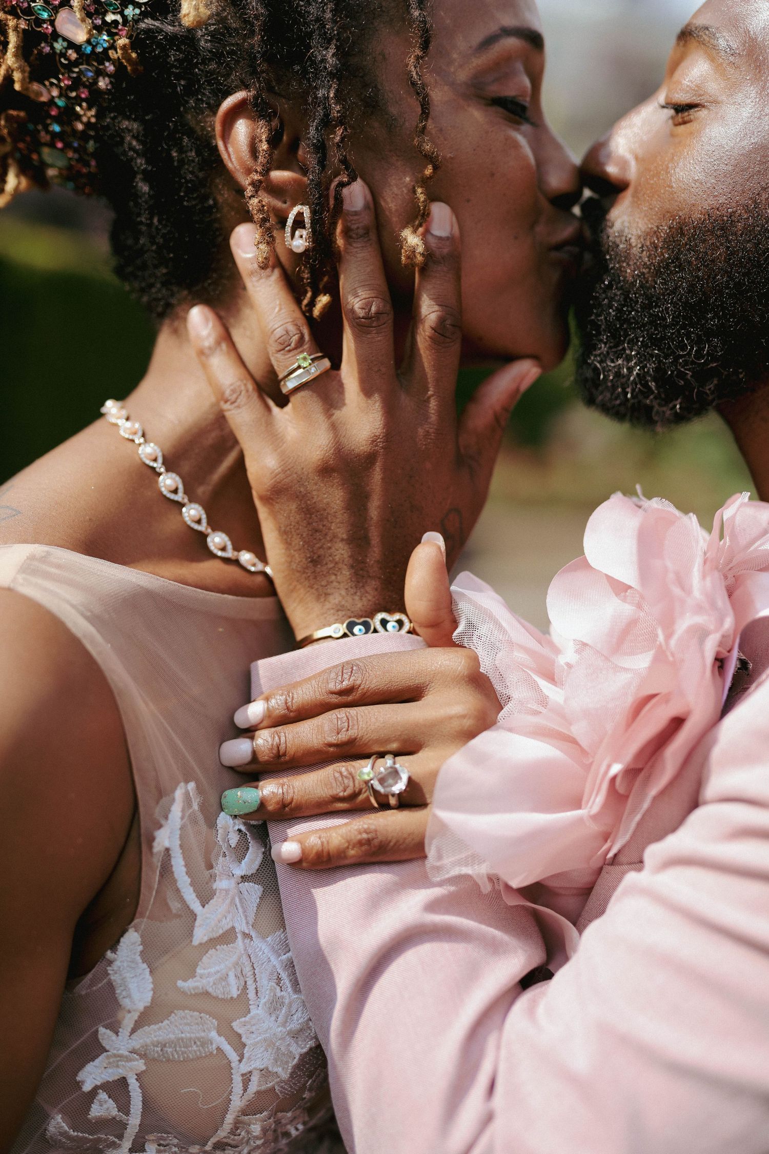 Couple sharing a close kiss, her hand cradling his bearded face, with stacked rings, a pearl necklace and a blush pink ruffled suit jacket.