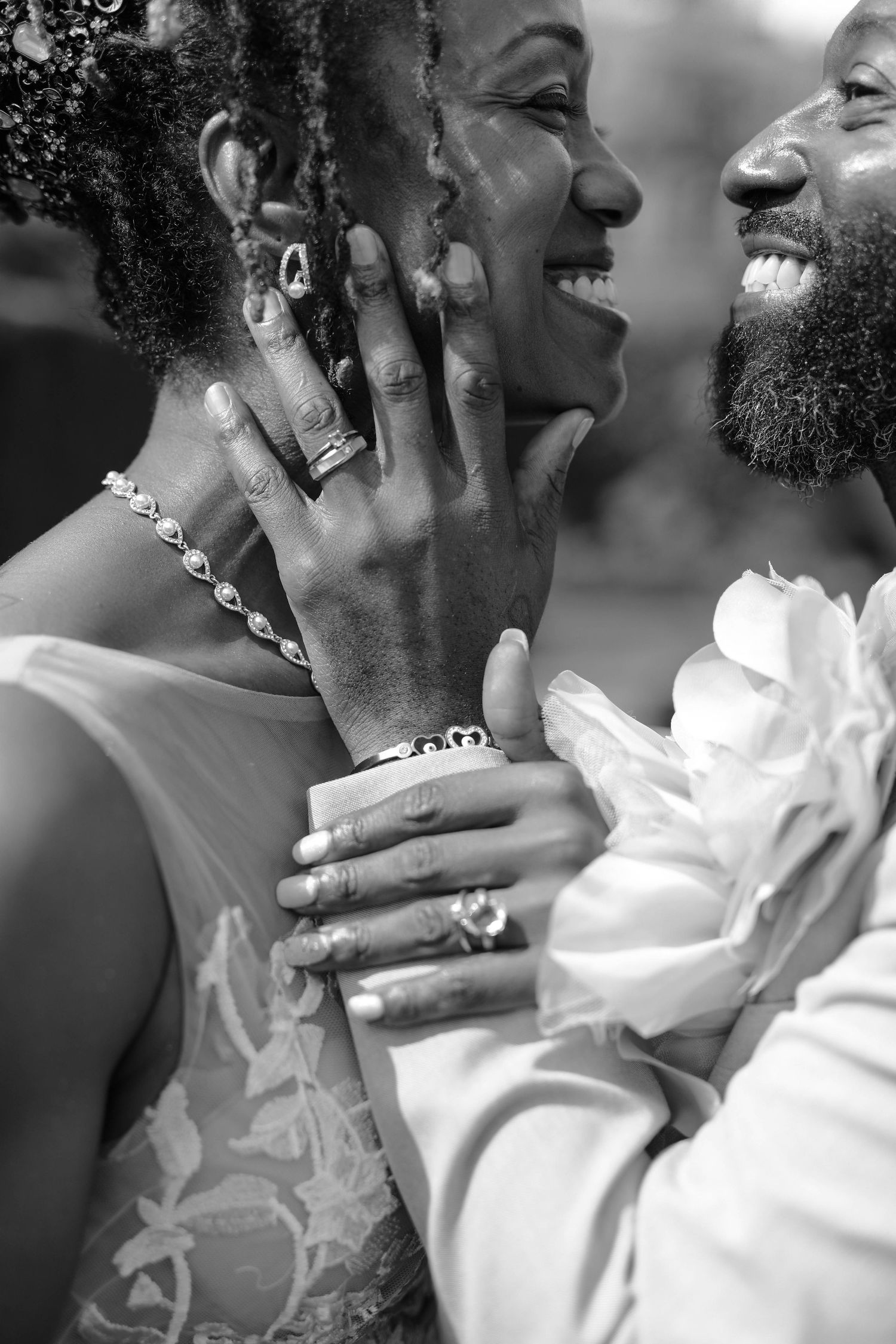 Black and white photo of a joyful couple laughing as she cups his face, wearing rings and a beaded necklace, in a tender wedding moment.