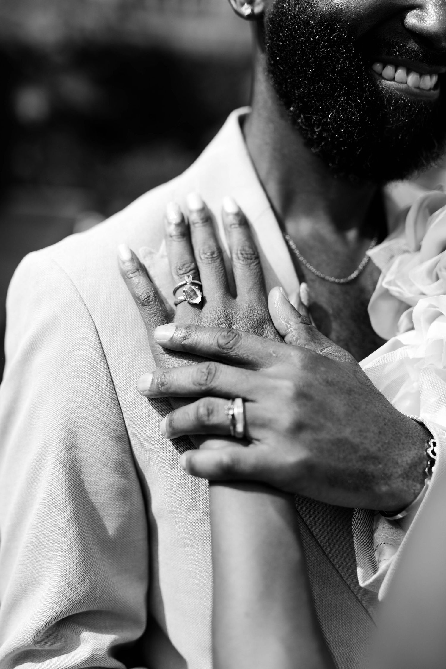 Black and white close-up of two hands with wedding rings resting on a smiling bearded man's chest, wearing a blazer and chain necklace.