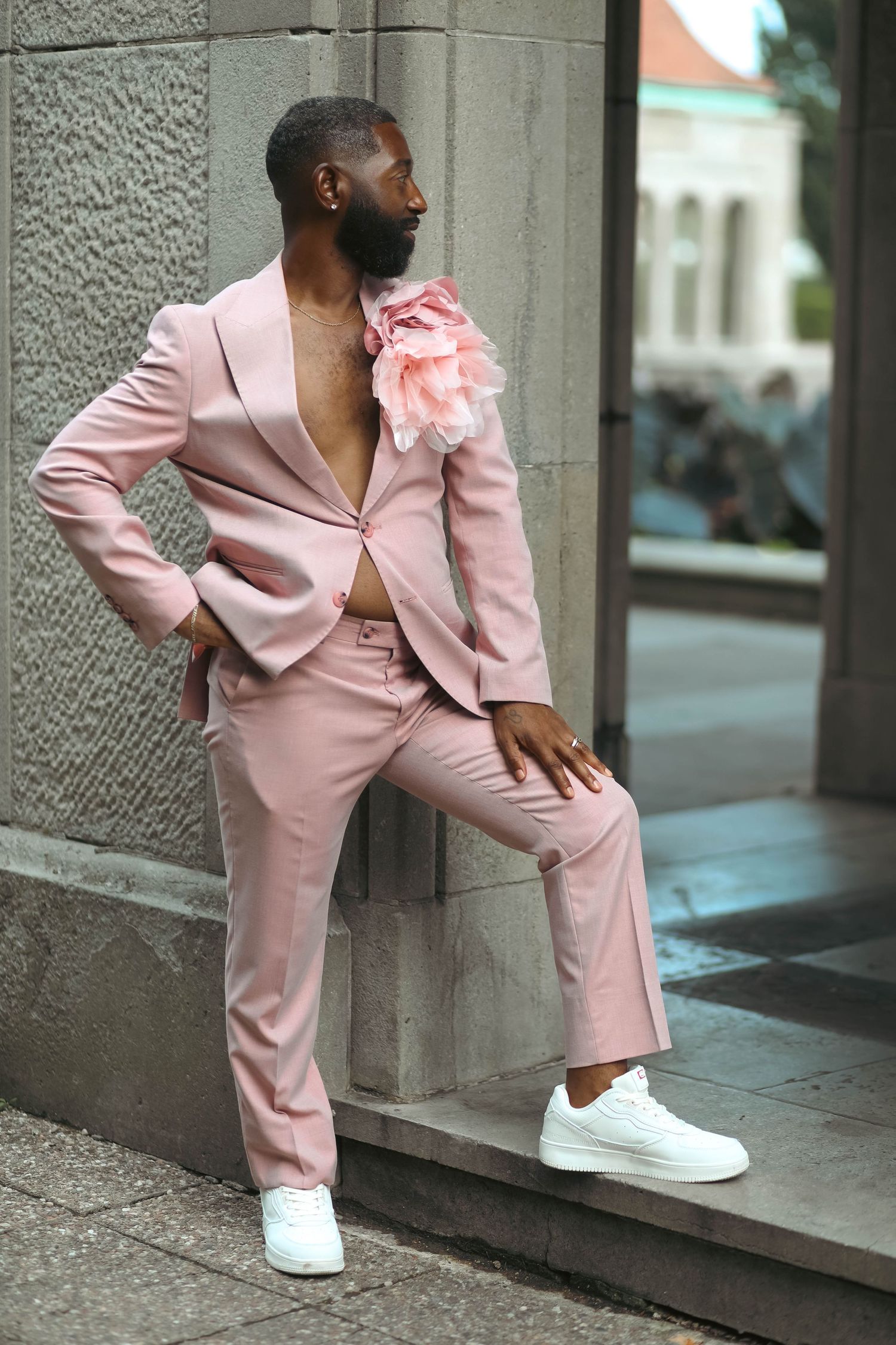 Man in a blush pink suit with a floral shoulder accent and white sneakers poses with one foot on a stone ledge.