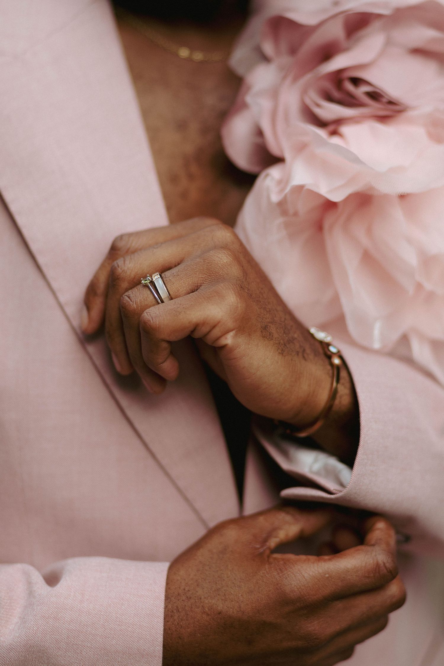 Close-up of hands adjusting a soft pink blazer, wearing a silver ring and gold bracelet, with a pink rose corsage on the lapel.