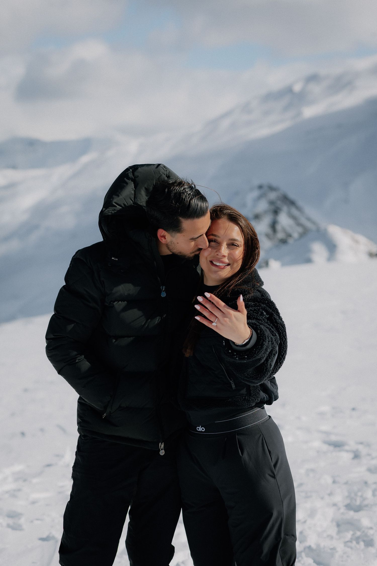 Couple in black outfits embracing on a snowy mountain summit, the woman showing her ring, with snow-covered peaks and clouds behind.