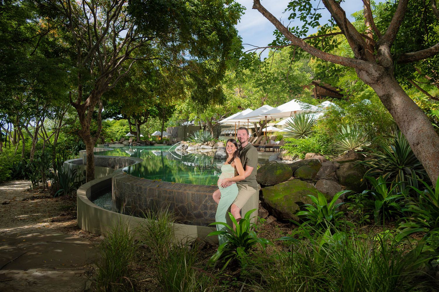 Couple embracing beside a stone-rimmed natural hot spring pool surrounded by lush tropical plants and tall trees at a scenic resort.