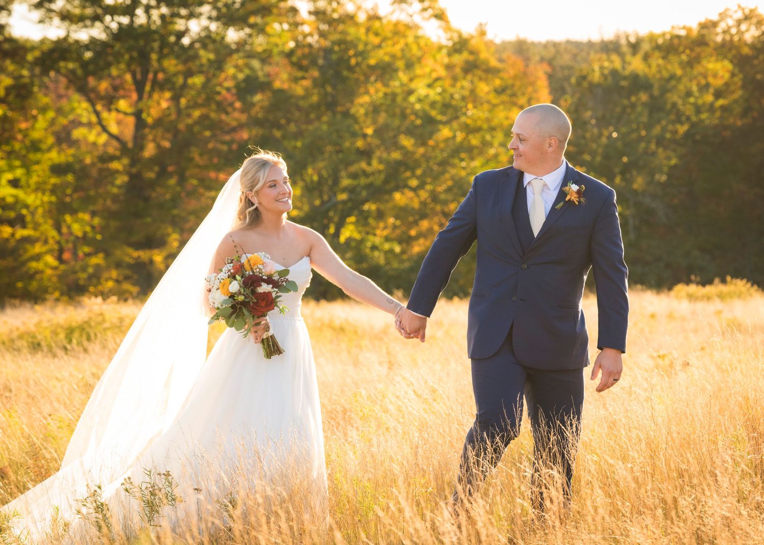 A smiling bride in a white gown and veil holds hands with her groom in a navy suit while walking through a golden field at sunset.
