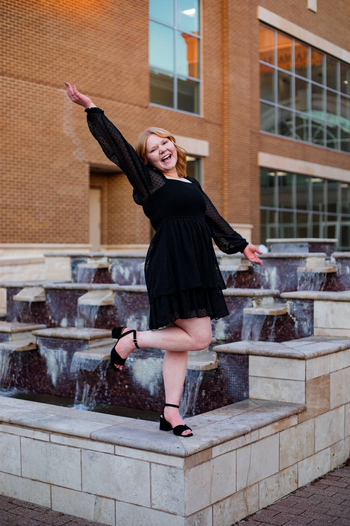 A joyful young woman in a black dress balances on one leg with her arm raised beside a fountain and brick building.