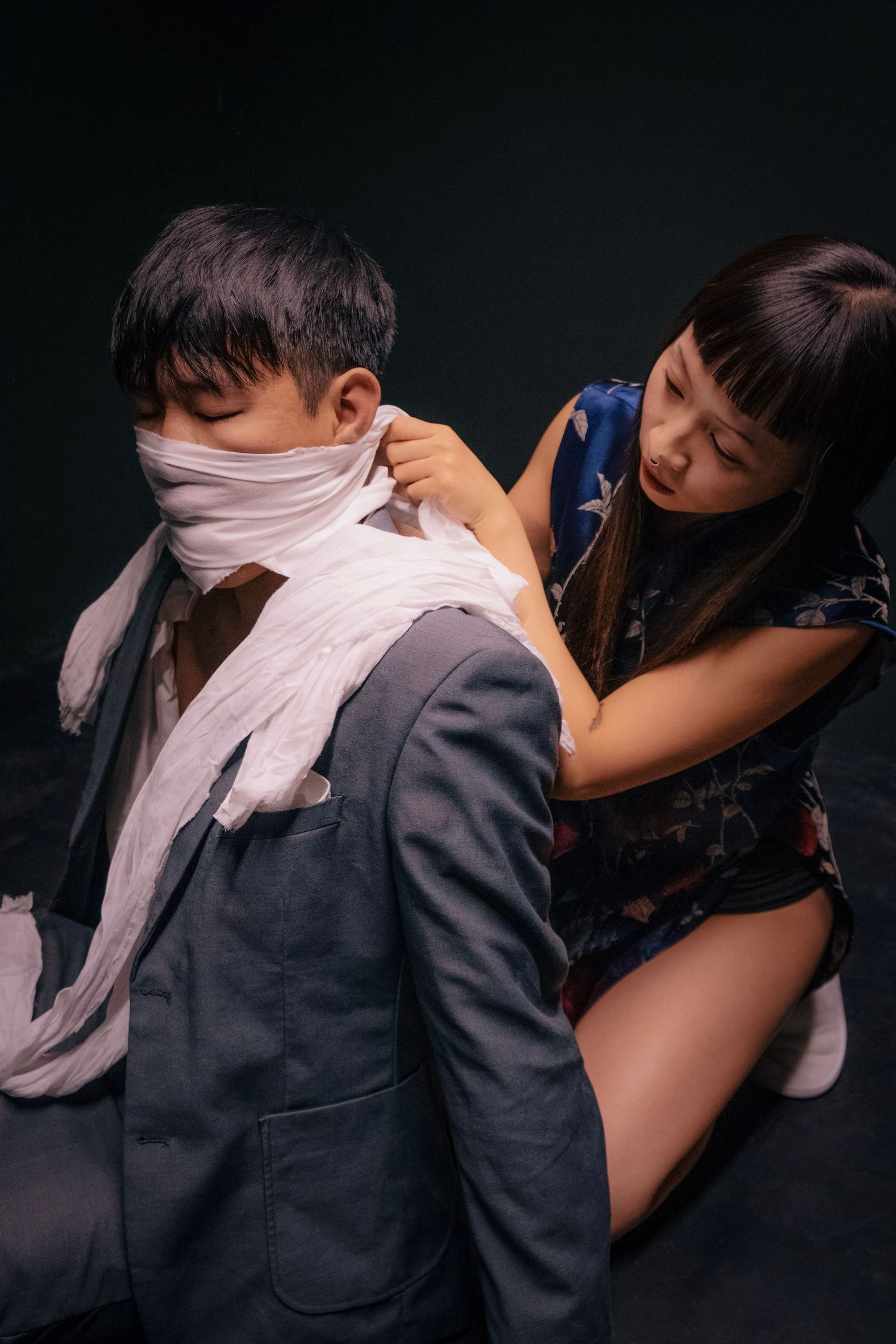 Woman wrapping white bandage around a man's face and neck in a dark studio setting.