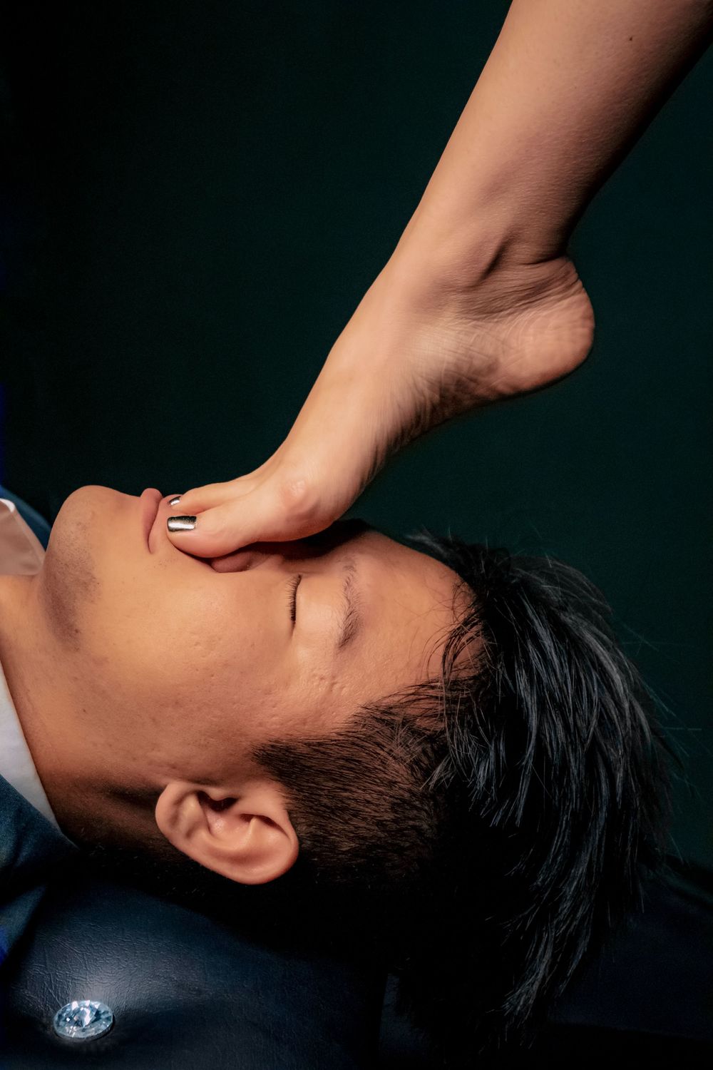 A woman's foot rests on a man's face as he lies back with eyes closed against a dark background.