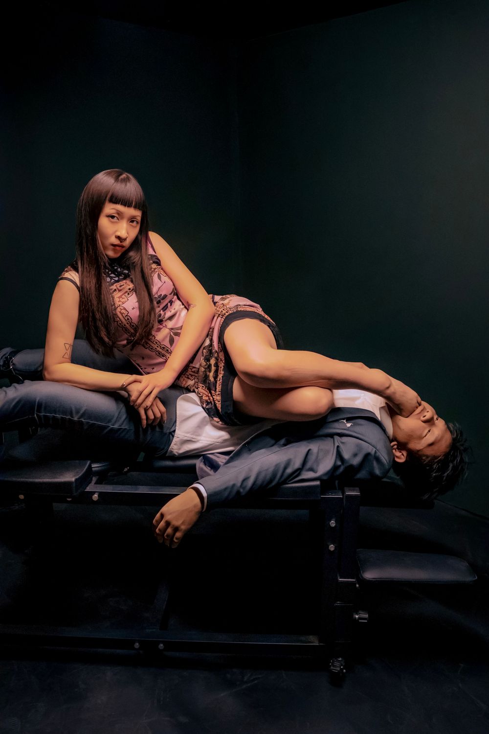 A woman in a floral dress sits on a bench while a man reclines across her lap in a dark studio setting.