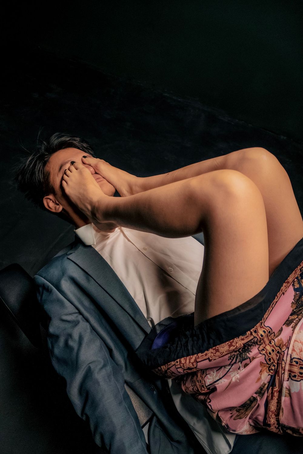A shirtless person reclines dramatically against dark background, wearing patterned skirt, covering face with hand.