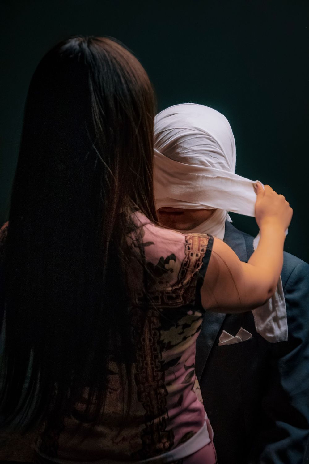 Woman with long dark hair wrapping white bandage around another person's head in a dark studio setting.