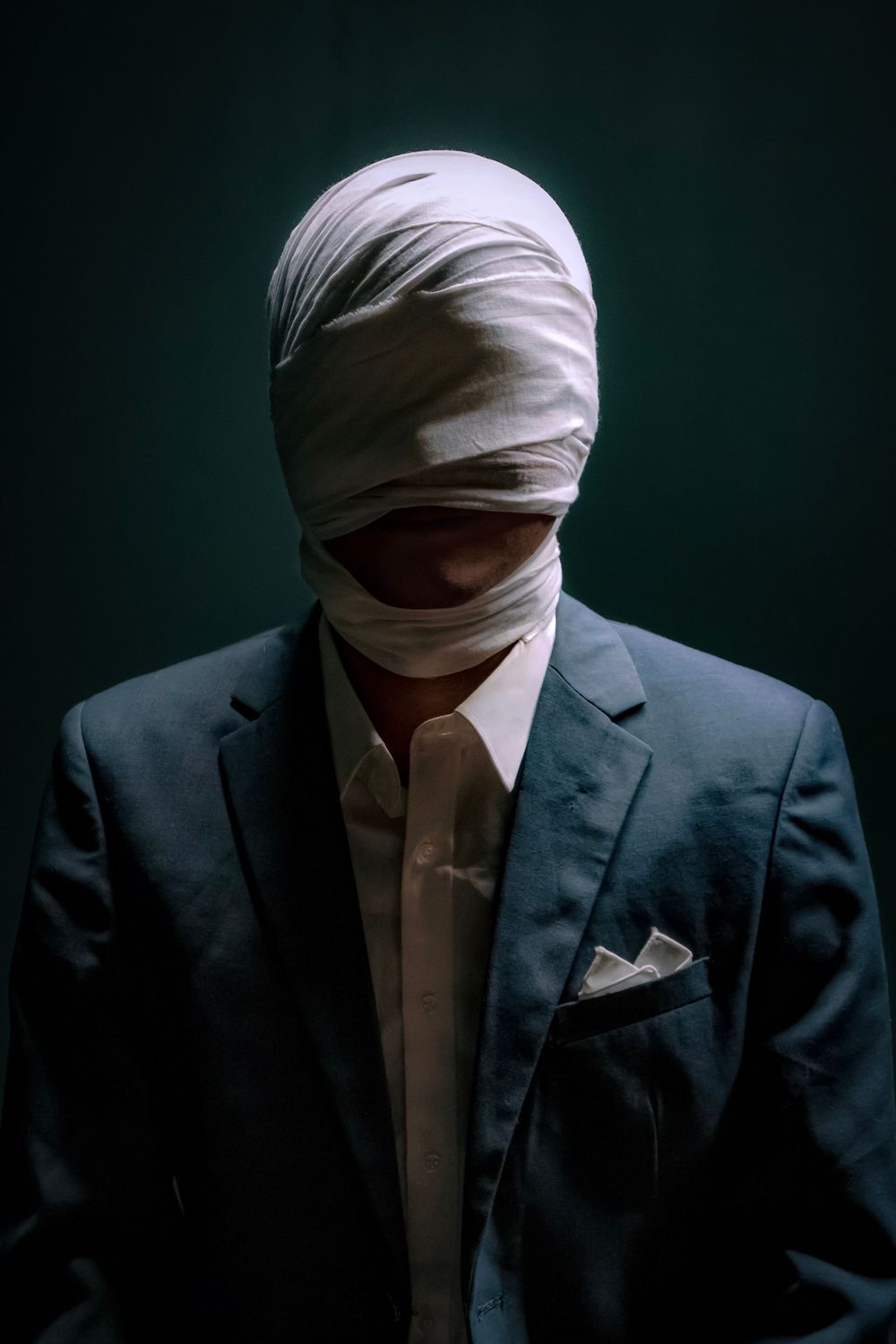A mysterious figure in a dark suit with their face wrapped in white bandages stands against a dark background.