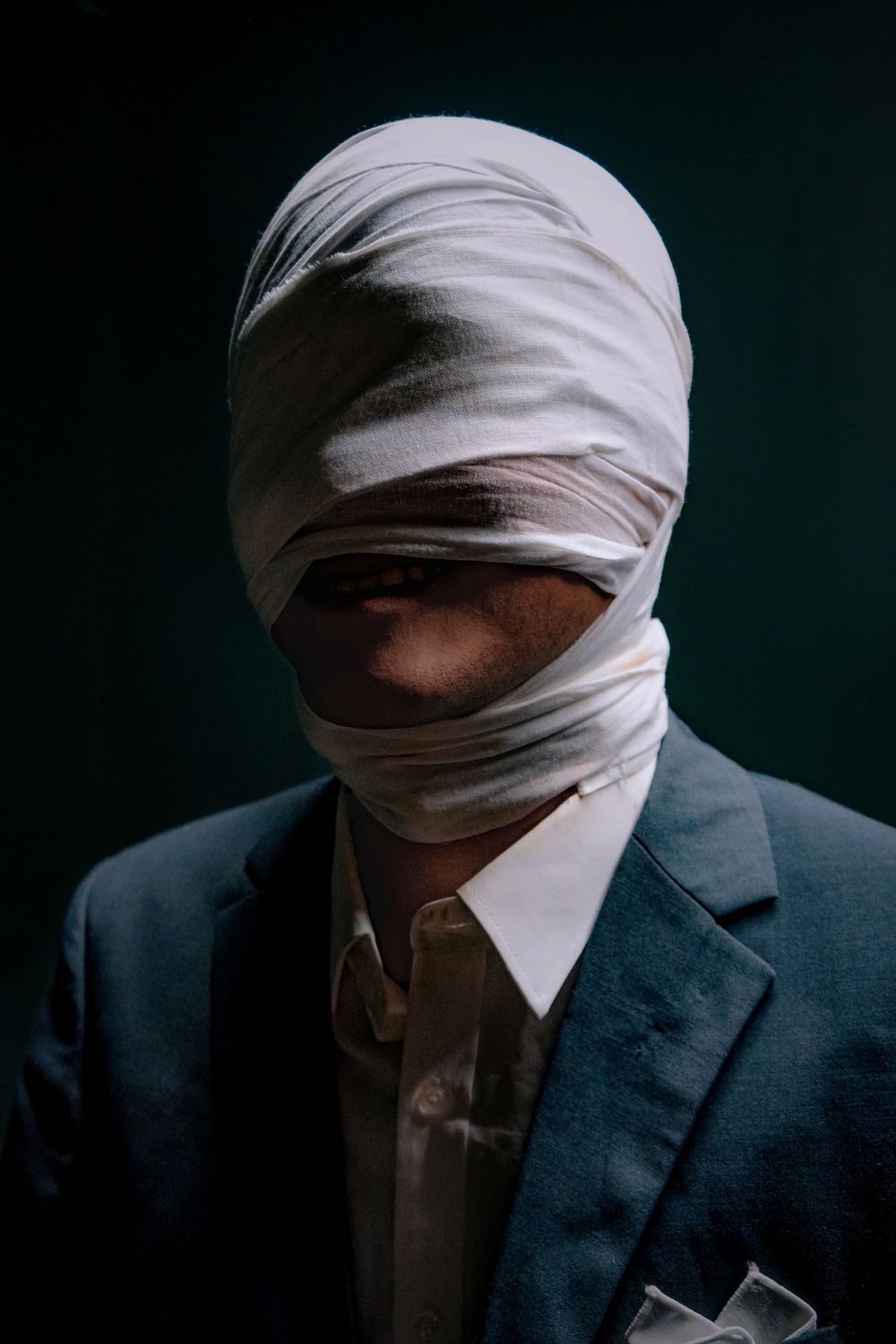 Man in blue suit with white bandages wrapped around his head against a dark background.