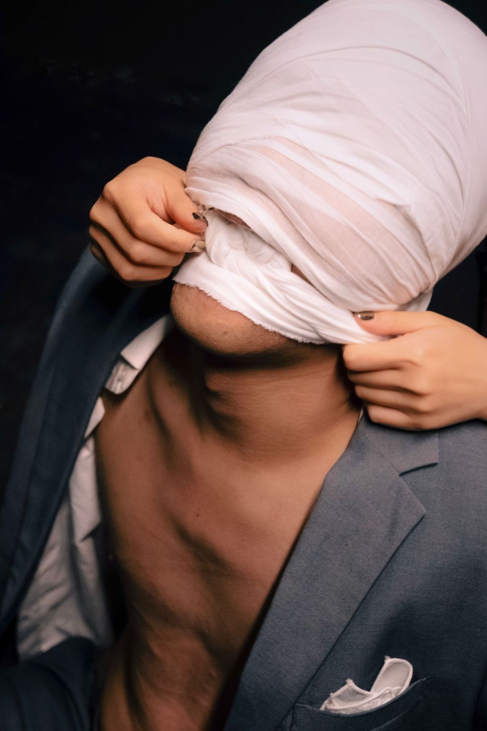 Man in gray suit removing white shirt over his head, revealing bare chest in a dark studio setting.