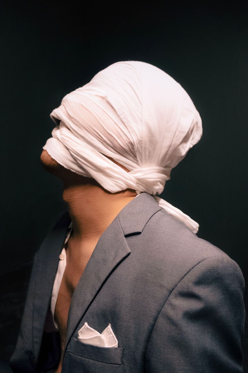 Person in gray suit with white fabric wrapped around their head, viewed from behind against dark background.