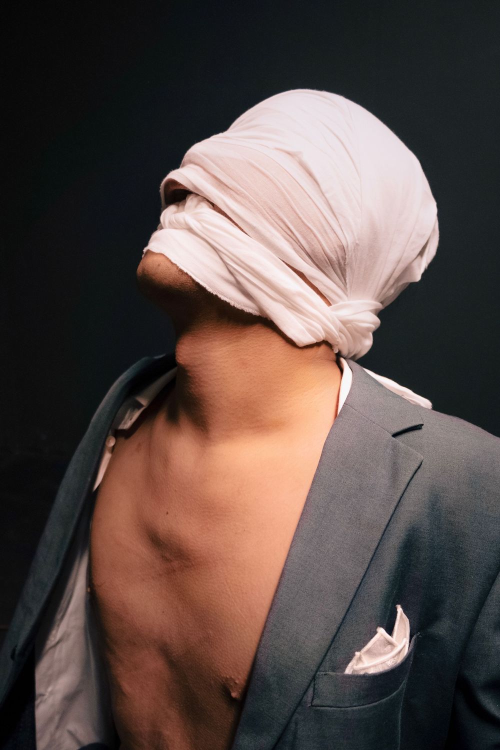 Person wearing a gray blazer with white cloth wrapped around their head against a dark background.