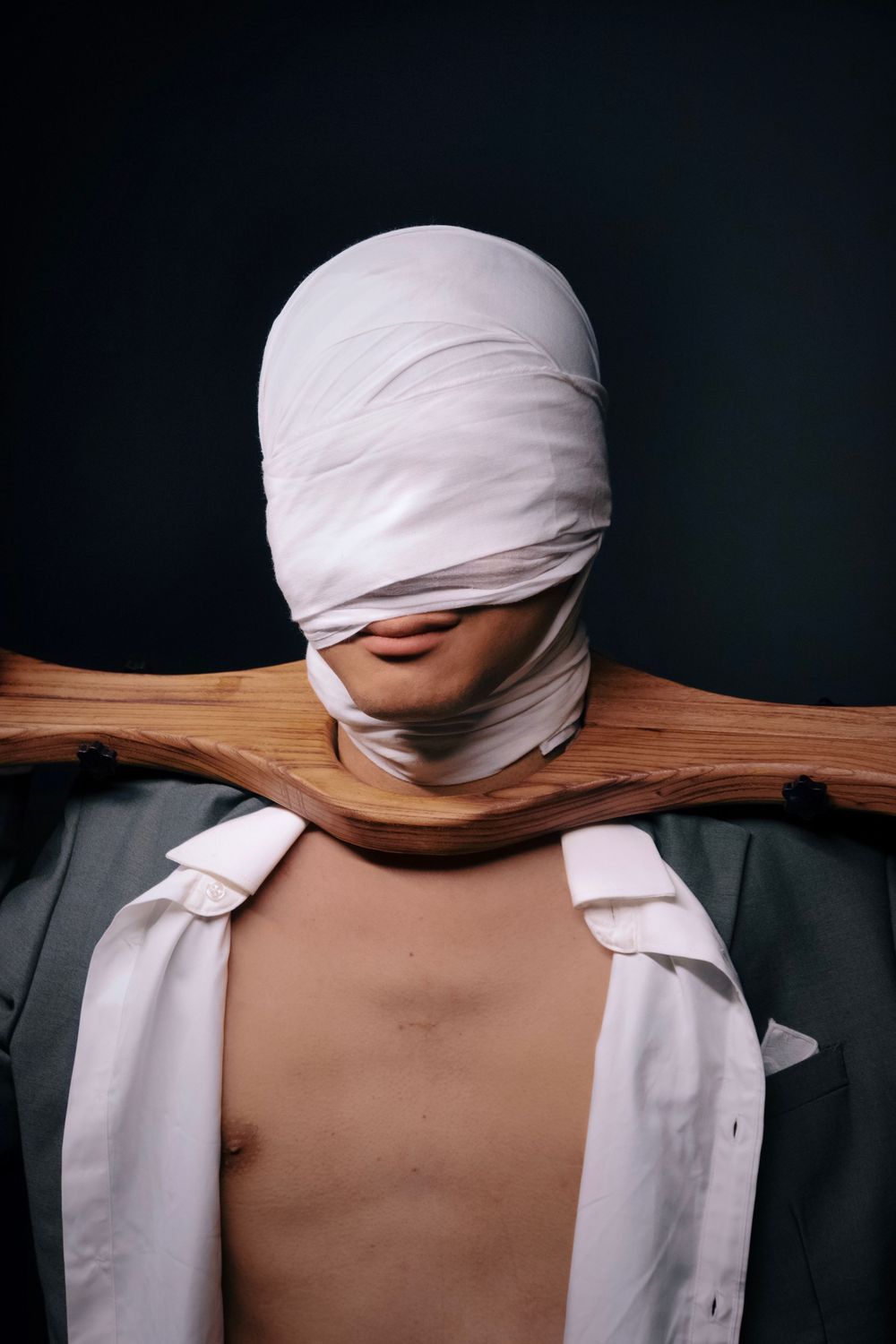 Person with white bandaged head held by wooden yoke, open shirt, dark background.